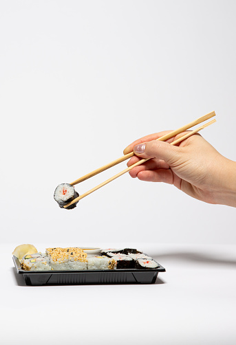 Woman's hand holding a sushi roll with wooden chopsticks, a black takeaway box isolated on white background