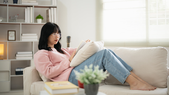 A woman is sitting on a couch with a book in her lap. She is wearing a pink sweater and blue jeans. The scene is cozy and comfortable, with a cup and a potted plant nearby