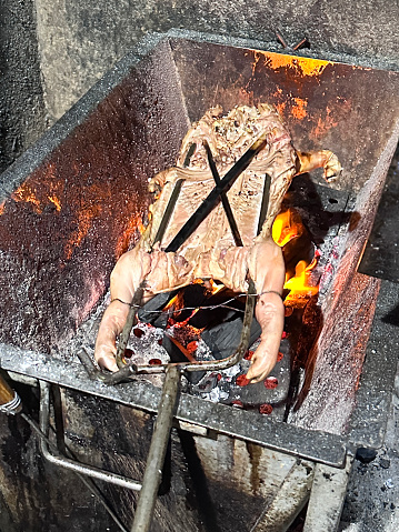 Stock photo showing close-up, elevated view of a piglet on a spit, lying on a metal rack over glowing, orange, flaming coals, that is being roasted to serve to customers at a Thailand back street food stall.