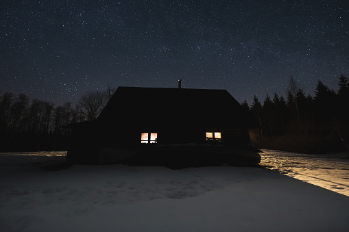 Night scene, landscape astrophoto. Silhouette of a house with light from a window in the forest against the background of the starry sky. Kollassaare farm in winter. High quality photo