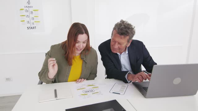young woman and a senior man are working together in a modern office, using a laptop and tablet to discuss a project laid out on paper in front of them