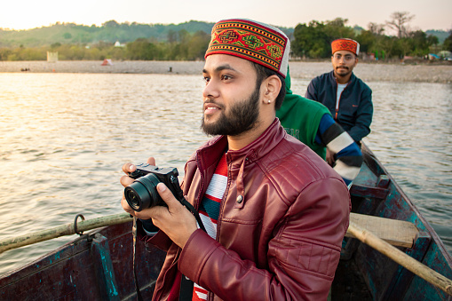 Portrait of young tourist, photographer man sitting on the sailboat and taking a photo of beautiful river view with the digital camera.  He Is wearing a typical cap of Himachal Pradesh.