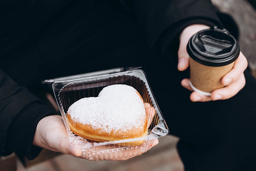 A paper cup of coffee and a heart-shaped donut in male hands, close-up. A man in a black jacket with a donut outside.