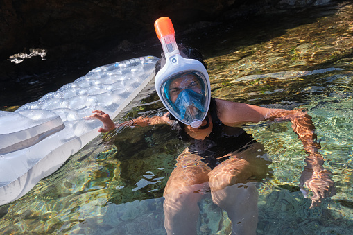 Female in swimwear and mask for snorkeling floating in clean ocean water with inflatable mattress and looking at camera