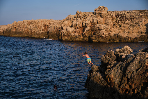 Unrecognizable child in swimwear jumping into ocean from edge of stone cliff enjoying summer holidays