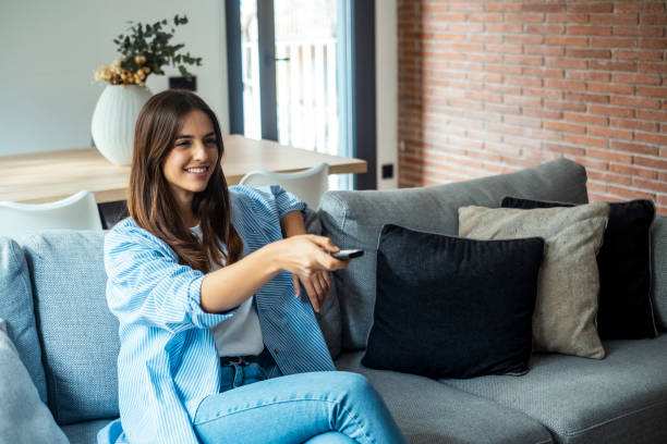 young woman sitting on her home sofa, holding the TV remote while enjoying a program. she is enjoying her leisure time at home. One female people using control tv watching movie alone on the couch