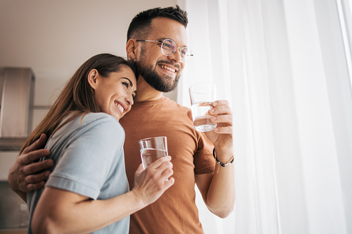 Photo of adult couple in the kitchen drinking water. They enjoy the view through the window.