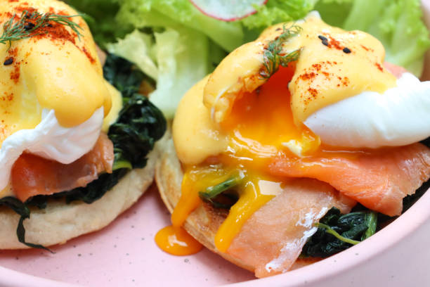 Stock photo showing a close-up view of a pink bow containing a breakfast of poached eggs with oozing yellow yolk, Eggs Benedict breakfast with smoked salmon and hollandaise sauce, topped with spinach and healthy salad.