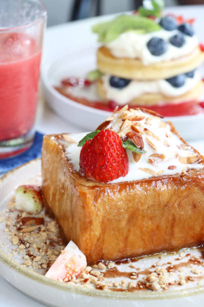 Close-up image of white plate of golden thick cut French toast with vanilla ice cream, strawberries and toasted almonds slices, American pancakes with whipped cream and fresh fruit, glass of strawberry and watermelon smoothie, al fresco dining scene