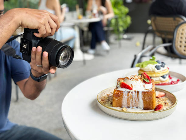 Image of unrecognisable photographer with DSL camera photographing white plate of golden thick cut French toast with vanilla ice cream, strawberries and toasted almonds slices, American pancakes with whipped cream and fresh fruit, al fresco dining scene