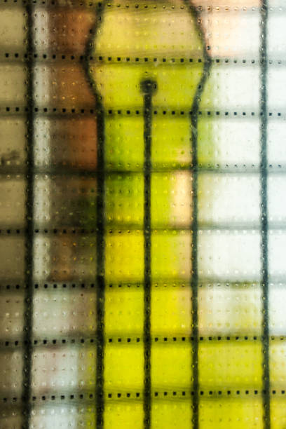 Close-up of a stained glass window with textured panels in blue, green, and clear tones. The grid-like surface diffuses the light, creating a soft and abstract effect. The image emphasizes geometry, color, and the artistic detail of architectural design.