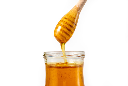Honey drips from a spoon into a jar on white background