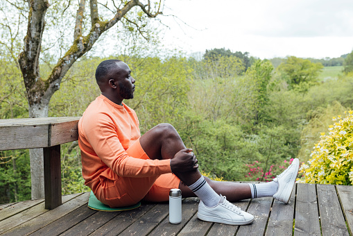 Man sitting on a wooden decking, looking out at a woodland view in Hexham, North East England. He is wearing active wear, taking a break from the exercise he has been doing.