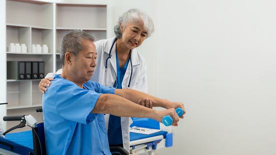 An elderly Asian man is doing physical therapy with the support of an elderly female nurse using dumbbells and tubes to exercise for a patient.