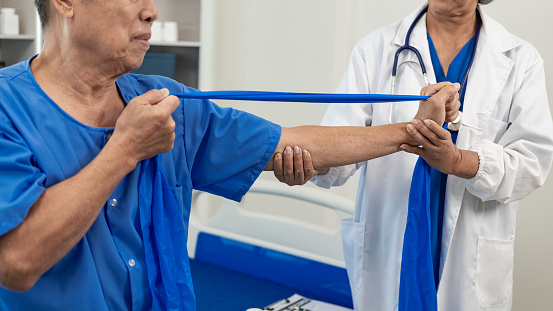 An elderly Asian man is doing physical therapy with the support of an elderly female nurse using dumbbells and tubes to exercise for a patient.