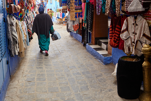 Blue city shopping street with Moroccan woman walking in traditional clothing in Chefchaouen, Morocco, North Africa.