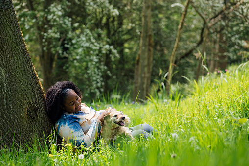 Wide shot view of a young woman taking a break, sitting down on the grass resting against a tree. She is in a woodland meadow with her dog outdoors in Hexham, North East England.