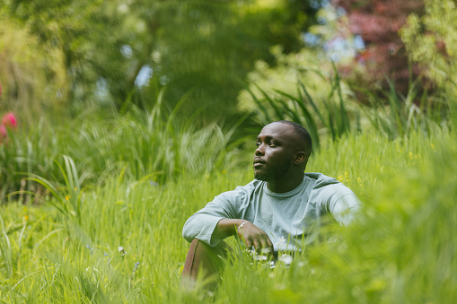 Man sitting in a long grass area in Hexham, North East England. He is looking away from the camera with a serious expression, practising mindfulness.