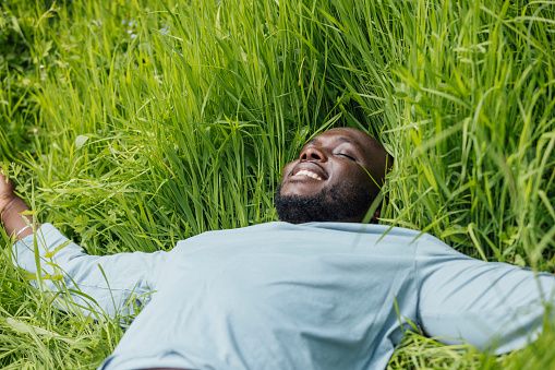 Medium shot of a man laying down with his arms outstretched in a long grass area in Hexham, North East England. He has his eyes closed and is smiling, practising mindfulness.