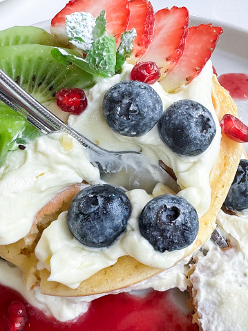 Stock photo showing elevated view of American-style, fluffy pancakes stack layered with whipped cream and fruit, topped with fresh blueberries, slices of kiwi and strawberry and drizzled with pomegranate seeds juice coulis, garnished with a mint sprig and served on a white plate.