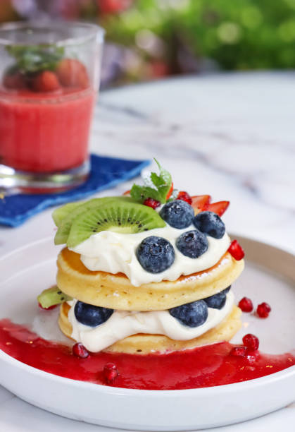 Stock photo showing close-up view of American-style, fluffy pancakes stack layered with whipped cream and fruit, topped with fresh blueberries, slices of kiwi and strawberry and drizzled with pomegranate seeds juice coulis, garnished with a mint sprig and served on a white plate.