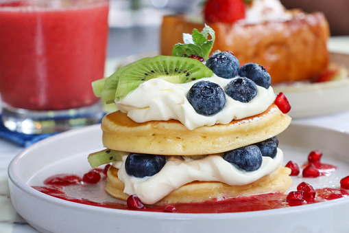 Stock photo showing close-up view of American-style, fluffy pancakes stack layered with whipped cream and fruit, topped with fresh blueberries, slices of kiwi and strawberry and drizzled with pomegranate seeds juice coulis, garnished with a mint sprig and served on a white plate.