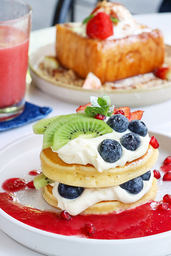 Stock photo showing close-up view of American-style, fluffy pancakes stack layered with whipped cream and fruit, topped with fresh blueberries, slices of kiwi and strawberry and drizzled with pomegranate seeds juice coulis, garnished with a mint sprig and served on a white plate.