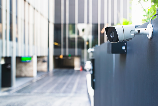 A modern security camera attached to a building's exterior wall, overseeing a blurred entrance area.