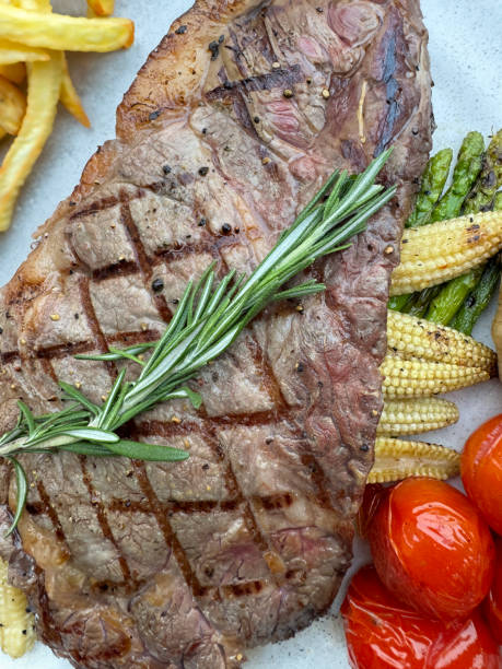istock Full frame image of white plate containing griddled sirloin steak with seared criss-cross lines and rosemary sprig, grilled tomatoes, French fries, asparagus, baby sweetcorn, elevated view 2068589655
