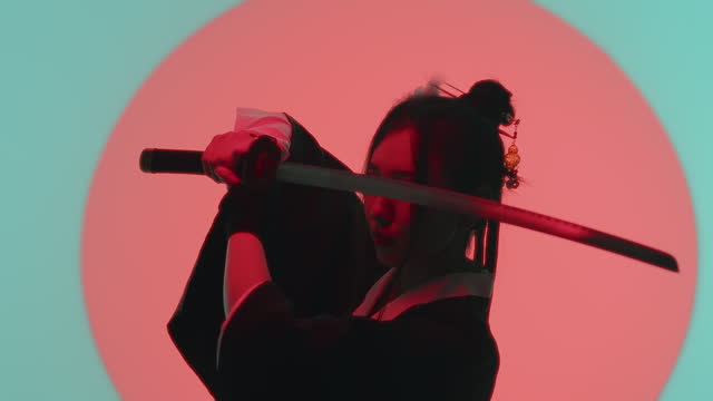 Young asian woman in a traditional kimono close up standing with a katana raised to eye level in the studio with a turquoise blue green background and red sun