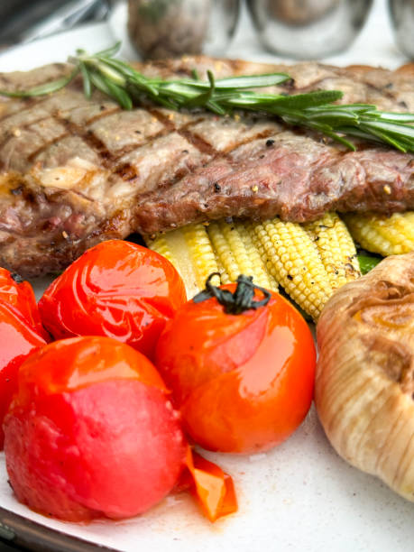 istock Full frame image of white plate containing griddled sirloin steak with seared criss-cross lines and rosemary sprig, grilled tomatoes, baby sweetcorn, roasted garlic bulb, metal cup background, elevated view 2068598093