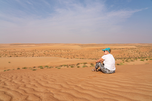 One mature women sitting in sand in Wahiba rippled desert in Oman in the morning.