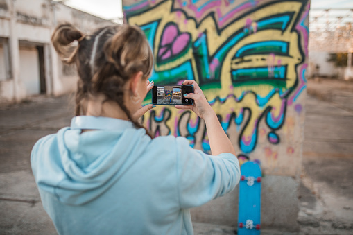 Back view of a young artist using a mobile phone to photograph the graffiti she drew on the wall of an abandoned building
