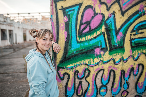 A smiling young artist looks directly into the camera as she stands by the wall of an abandoned building where she has drawn graffiti