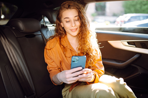 Young woman uses a smartphone while sitting in the back seat of a car. Concept of technology, traveling by car, business.