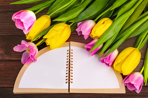 Open notebook with space for text and bouquet of pink and yellow tulip flowers around on dark wooden background.