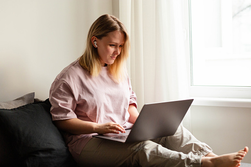 Blonde woman in a pink t-shirt working from home on a couch by the window with a laptop looking at monitor with wireless headphones