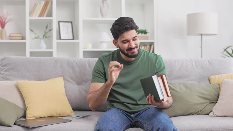 Indian man with books on couch showing on sign with fingers