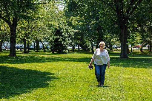 A mature woman with short blond hair, wearing glasses, and carrying shoes in her hand, enjoys a leisurely walk on the lush green grass of a sunlit park in Washington, D.C.