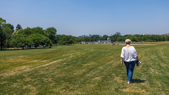 A mature Caucasian blonde woman in casual clothes, is walking on the green grass of the National Mall on a sunny day in Washington, D.C. Lincoln Memorial shown in the distance