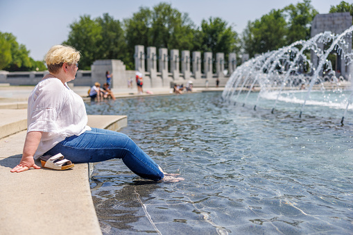 Caucasian mature blonde woman in glasses, sitting with feet in fountain by memorial. The World War II Memorial towers the Rainbow Pool in Washington, D.C.