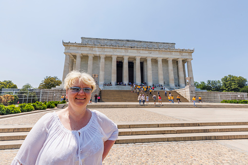 A smiling Caucasian blonde female tourist in a white blouse, stands in front of the Lincoln Memorial, a popular historical landmark in Washington, D.C. The clear blue sky and bright summer day
