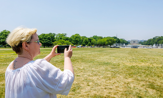 A mature blonde woman with a tattoo, captures the World War II Memorial using her smartphone, with the Lincoln Memorial Reflecting Pool in the far distance in Washington, D.C.