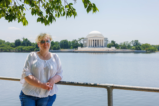 A mature woman with blonde hair and sunglasses stands in front of the Jefferson Memorial across the Tidal Basin in Washington, D.C.