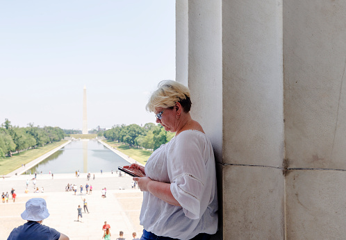 A mature Caucasian blonde woman taking a break at shadow of Lincoln Memorial in Washington, D.C. Clear blue sky and the Reflecting Pool, with tourists, visible in the distance