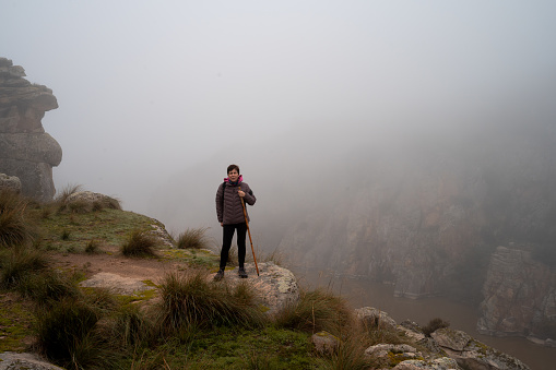 From the edge of a cliff, at the Douro river crossing, a woman in hiking clothes observes the landscape with low visibility due to the dense fog. Hiking activity in autumn, a cold morning with wet fog.