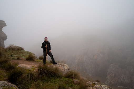 From the edge of a cliff, with the Douro River in the background, a man in trekking clothes observes the landscape with low visibility due to the dense fog. Hiking activity in autumn, a cold morning with wet fog.