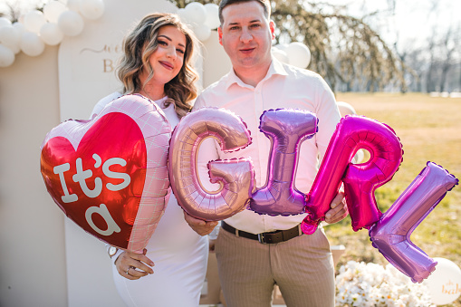 Smiling parents are standing in the park and holding balloons that say: It's a girl!