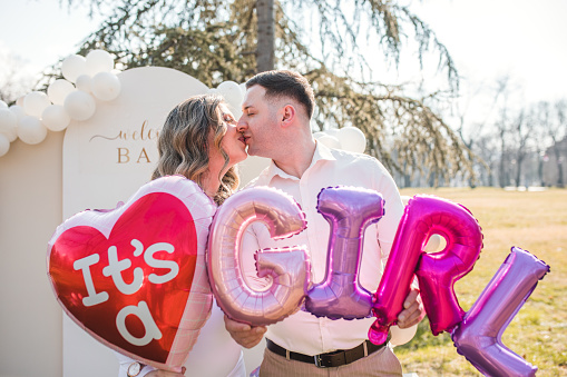 Future parents are standing in the park and holding balloons that say: It's a girl!