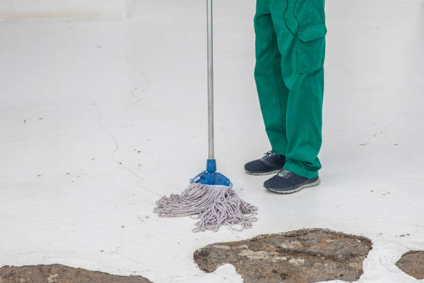 Cleaner wearing green overalls using a mop to clean a bright white floor.